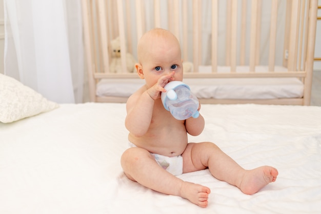 8-miesięczny chłopiec siedzi w pieluchach na białym łóżku z butelką mleka w domu, koncepcja jedzenia dla niemowląt, pierwsza przynęta