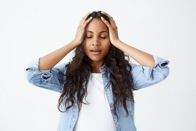 Afro-amerykańska studentka o długich falowanych włosach od niechcenia ubrana czuje się zestresowana, trzyma ręce na głowie, z zamkniętymi oczami w frustracji i rozpaczy po tym, jak popełniła poważny błąd.