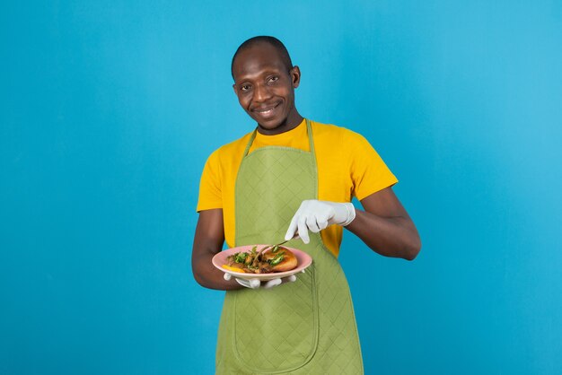 Afro amerykański mężczyzna w zielonym fartuchu trzyma talerz z jedzeniem na niebieskiej ścianie