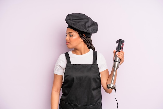 Afro czarna kobieta o widoku profilu myśląca, wyobrażająca sobie lub marząca. szef kuchni z blenderem ręcznym