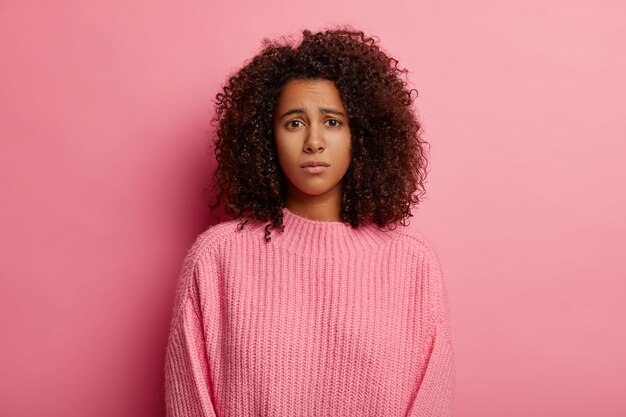 Afro kobieta z żałosnym spojrzeniem, niezadowoloną miną, ubrana w zwykłe ciuchy, niezadowolona ze złych wieści, smutno patrzy w kamerę, nosi sweter, odizolowana na różowym tle.