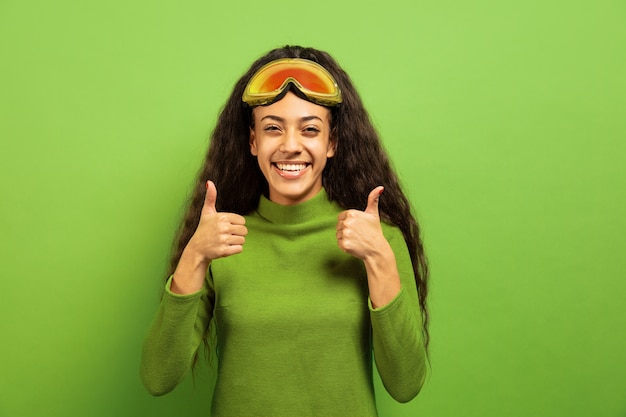 Afro-portret młodej kobiety brunetka w masce narciarskiej na tle zielonym studio. Pojęcie ludzkich emocji, wyrazu twarzy, sprzedaży, reklamy, sportów zimowych i wakacji. Uśmiechnięty, kciuk do góry.