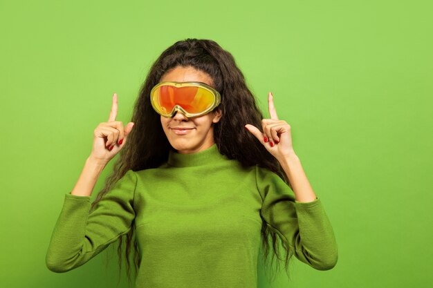 Afro-portret młodej kobiety brunetka w masce narciarskiej na tle zielonym studio. Pojęcie ludzkich emocji, wyrazu twarzy, sprzedaży, reklamy, sportów zimowych i wakacji. Uśmiechnięty, wskazujący w górę.