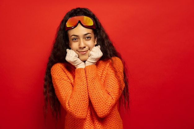 Afro-portret młodej kobiety brunetka w maska narciarska na tle czerwonym studio. Pojęcie ludzkich emocji, wyrazu twarzy, sprzedaży, reklamy, sportów zimowych i wakacji. Uśmiechnięty, wygląda uroczo.