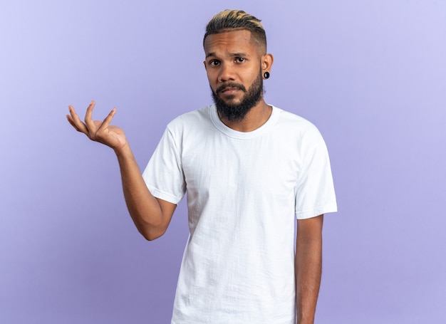 Afroamerykanin, młody człowiek w białej koszulce, patrząc na kamerę z mylącym wyrazem, podnosząc rękę w oburzeniu stojąc na niebieskim tle