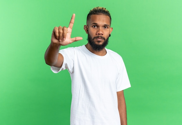 Afroamerykanin młody człowiek w białej koszulce patrzący na kamerę z poważną twarzą pokazującą palec wskazujący stojący nad zielonym tłem