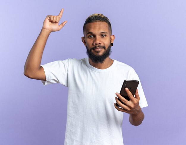 Afroamerykanin, młody człowiek w białej koszulce, trzymając smartfon pokazujący palec wskazujący, patrząc na kamery szczęśliwy i pewny siebie nowy pomysł koncepcji