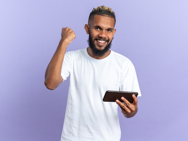 Afroamerykanin, młody człowiek w białej koszulce, trzymając smartfona, zaciskając pięść, szczęśliwy i podekscytowany, ciesząc się jego sukcesem, stojąc na niebieskim tle