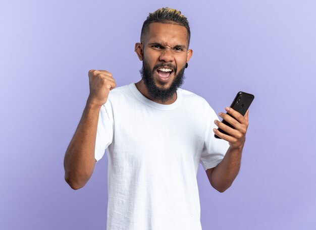 Afroamerykanin, młody człowiek w białej koszulce, trzymając smartfona, zaciskając pięść, szczęśliwy i podekscytowany, krzycząc ciesząc się swoim sukcesem, stojąc na niebieskim tle