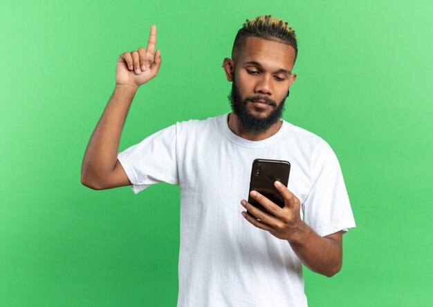 Afroamerykanin, młody człowiek w białej koszulce, trzymający smartfona, patrzący na niego pokazujący palec wskazujący mający nowy pomysł stojący nad zielonym tłem