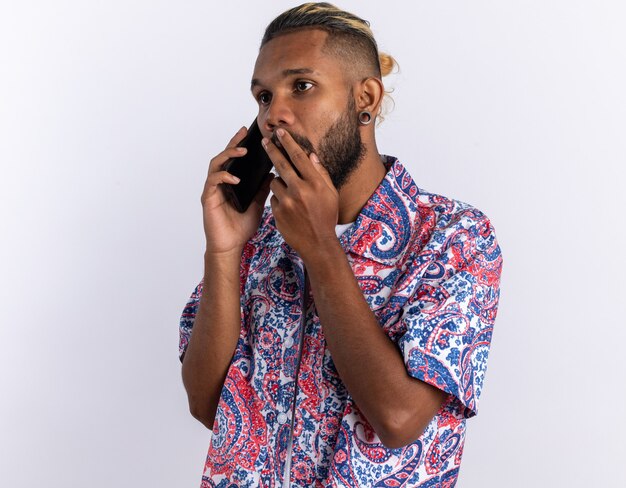 Afroamerykanin młody człowiek w kolorowej koszuli wyglądający na zdziwionego podczas rozmowy przez telefon komórkowy stojący nad białymi