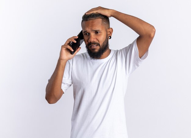Afroamerykanin młody mężczyzna w białej koszulce wyglądający na zdezorientowanego podczas rozmowy przez telefon komórkowy stojący nad białymi