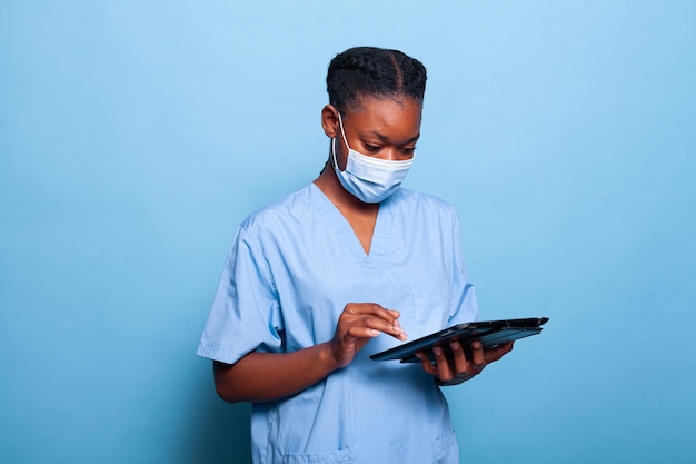 Afroamerykanin praktyk pielęgniarka z medyczną maską na twarz przeciwko koronawirusowi wpisując wiedzę o chorobie na komputerze typu tablet pracujący w leczeniu w studio. usługi medyczne