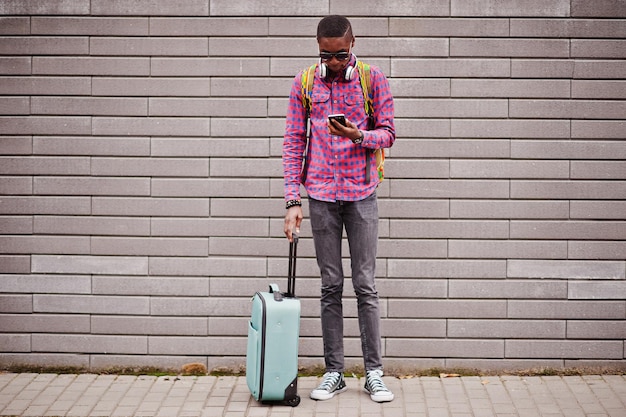Afroamerykanin w okularach przeciwsłonecznych w kratkę i słuchawkach z walizką i plecakiem Czarny podróżnik na ścianie trzymający telefon komórkowy