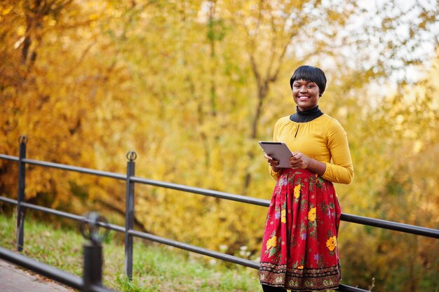 Afroamerykanka w żółtej i czerwonej sukience w złotym jesiennym parku z tabletem na rękach