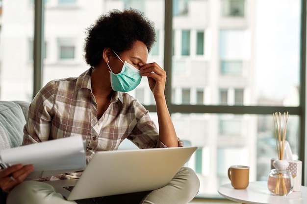 Bezpłatne zdjęcie afroamerykanka z maską na twarz ma ból głowy podczas wypełniania formalności i korzystania z laptopa podczas pracy w domu