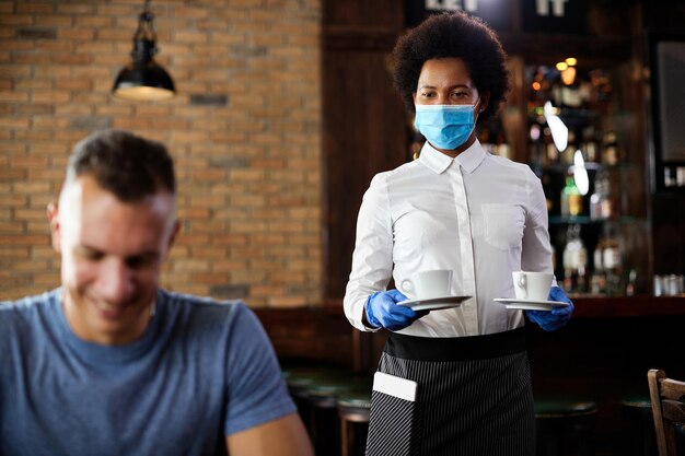 Afroamerykańska kelnerka serwująca kawę w masce i rękawiczkach