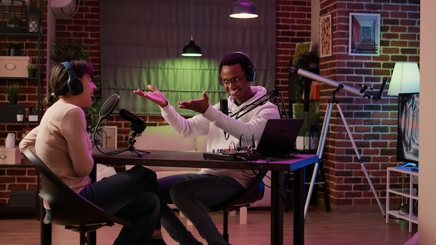 Bezpłatne zdjęcie afroamerykański podcaster wprowadzający gościa do słuchaczy w wieczornym programie online transmitowanym z domowego studia. zróżnicowana para rozmawia w internetowym wywiadzie na żywo przy użyciu profesjonalnych narzędzi audio.