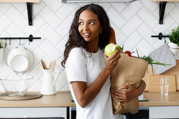 Afrykańska dziewczyna stojąca w kuchni trzyma papierową torbę z jedzeniem i je jabłko