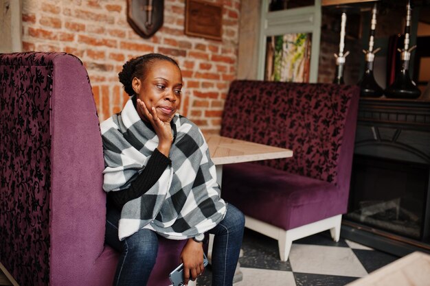 Afrykańska kobieta w kraciastej pelerynie pozowała w kawiarni z telefonem komórkowym w rękach