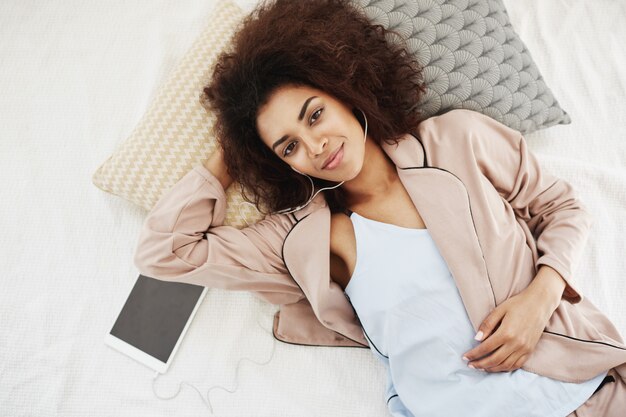 Bezpłatne zdjęcie afrykańska kobieta w piżamie uśmiecha się słuchanie muzyki w słuchawkach leżąc na łóżku.