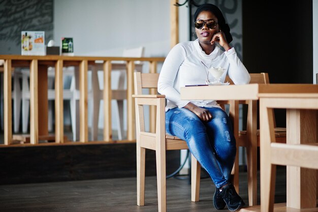 Afrykańska muzułmanka w czarnym hidżabie i okularach przeciwsłonecznych siedzi w kawiarni i je lody