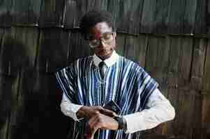 Bezpłatne zdjęcie afrykański biznesmen w tradycyjnych ubraniach i okularach z telefonem komórkowym pod ręką, patrząc na swoje zegarki na drewnianej ścianie