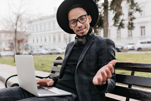 Afrykański freelancer wyrażający zdumienie podczas pracy z laptopem w kwadracie. Odkryte zdjęcie zaskoczonego czarnego mężczyzny w kapeluszu, siedzącego na ławce i trzymającego komputer.