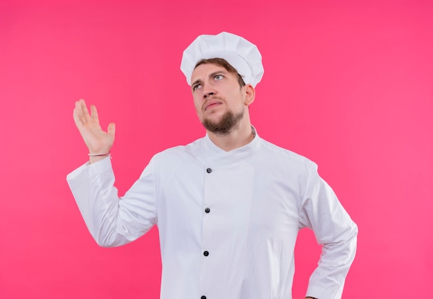Agresywny młody brodaty szef kuchni w białym mundurze, podnosząc rękę i myśląc na różowej ścianie