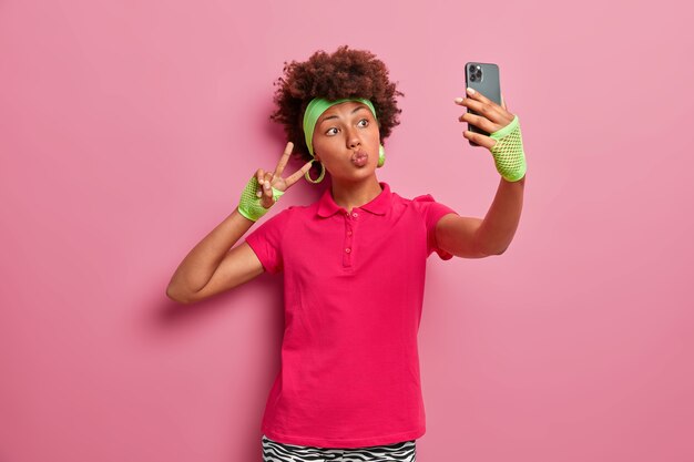 Aktywna kobieta z kręconymi włosami w różowej koszulce, opasce na głowę i sportowych rękawiczkach, robi selfie, wykonuje gest zwycięstwa, trzyma telefon komórkowy, ma obsesję na punkcie sieci społecznościowych, pozuje w domu