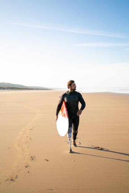 Aktywny surfer z amputowaną nogą na plaży z deską surfingową. Brodata osoba po amputacji w kombinezonie chodzącym po piasku, niosąca deskę i odwracająca wzrok