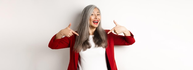 Bezpłatne zdjęcie ambitna starsza kobieta wskazująca palce na centrum, pokazująca logo i uśmiechająca się stojąca nad białym backg