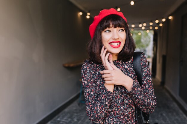 Animowana brunetka w czerwonym berecie i sukience vintage ociera ręce, postanawiając wyśmiać przyjaciela. Szczegół portret podekscytowany młoda kobieta z ciemnymi włosami krótkimi i uśmiechem hollywood, odwracając