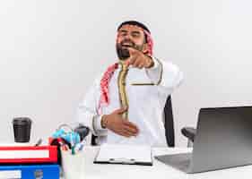 Bezpłatne zdjęcie arabski biznesmen w tradycyjnej odzieży siedzi przy stole z laptopem śmiejąc się wskazując palcem do ciebie, pracującego w biurze