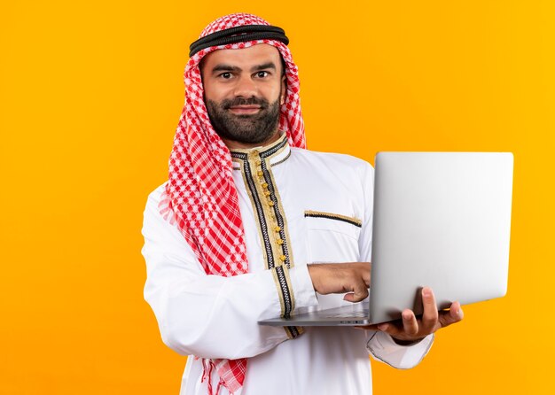 Arabski biznesmen w tradycyjnym nosić trzymając laptopa z uśmiechem na twarzy stojącej nad pomarańczową ścianą
