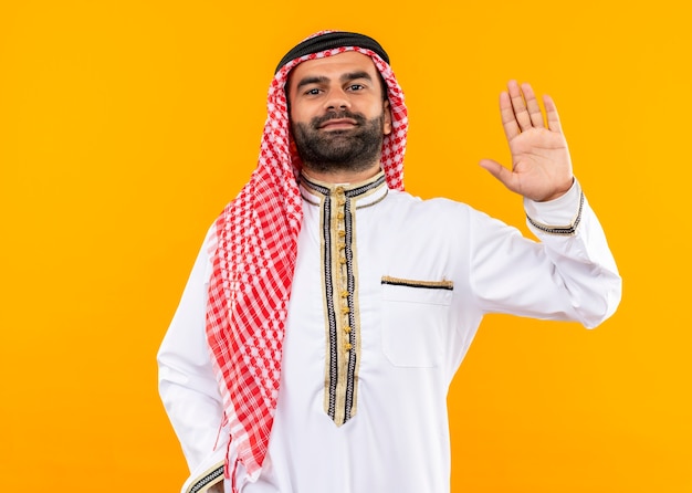 Arabski biznesmen w tradycyjnym stroju, pewnie macha ręką stojącą na pomarańczowej ścianie
