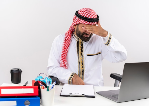 Arabski biznesmen w tradycyjnym stroju siedzi przy stole z laptopem patrząc zmęczony i znudzony zasłaniając oczy ręką pracującą w biurze