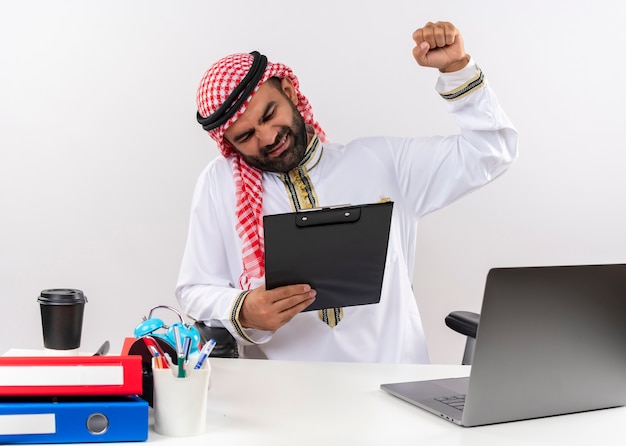 Arabski Biznesmen W Tradycyjnym Stroju Siedzi Przy Stole Z Laptopem Trzymając Schowek Zaciskający Pięść Z Zirytowaną Miną Pracujący W Biurze