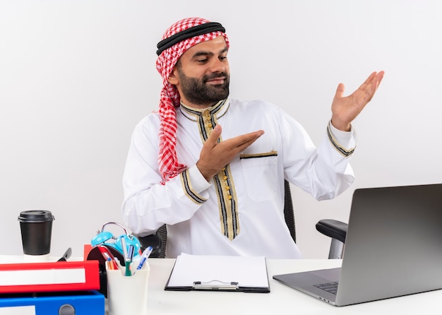 Bezpłatne zdjęcie arabski biznesmen w tradycyjnym stroju siedzi przy stole z laptopem, wskazując z rękami na bok, patrząc pewnie pracując w biurze