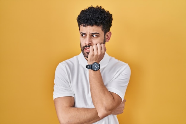 Arabski mężczyzna stojący na żółtym tle, wyglądający na zestresowanego i zdenerwowanego z rękami na ustach obgryzającymi paznokcie. problem z lękiem.