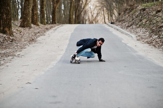 Arabski mężczyzna w stylu ulicznym w okularach z longboardowym longboardem w dół drogi