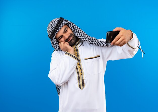 Arabski mężczyzna w tradycyjnym stroju, biorąc selfie za pomocą smartfona, uśmiechając się ze szczęśliwą twarzą stojącą nad niebieską ścianą
