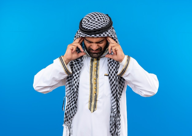Bezpłatne zdjęcie arabski mężczyzna w tradycyjnym stroju, dotykający swoich skroni, źle wyglądający cierpiący na silny ból głowy, stojący nad niebieską ścianą