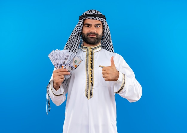 Arabski mężczyzna w tradycyjnym stroju pokazując gotówkę uśmiechając się pewnie pokazując kciuki do góry stojąc na niebieskiej ścianie