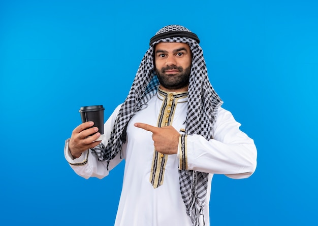 Arabski mężczyzna w tradycyjnym stroju pokazuje smartfon wskazujący palcem na niego z uśmiechem na twarzy stojącej nad niebieską ścianą