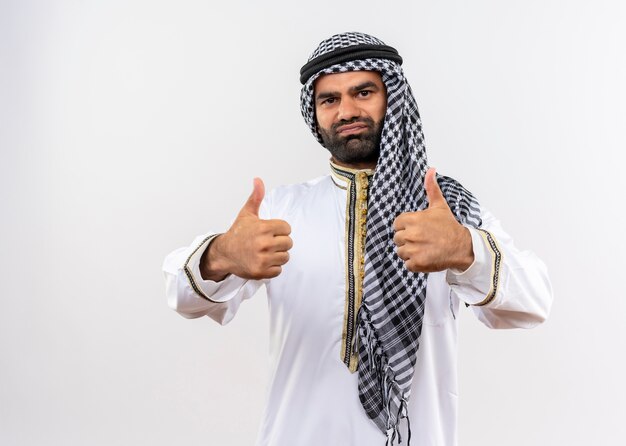 Arabski mężczyzna w tradycyjnym stroju uśmiechnięty pokazując kciuki stojąc na białej ścianie