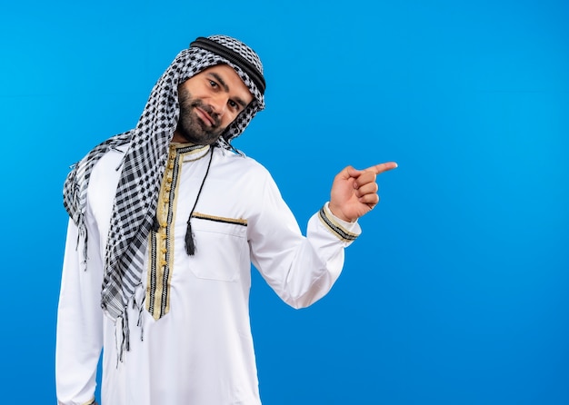 Bezpłatne zdjęcie arabski mężczyzna w tradycyjnym stroju z uśmiechem na twarzy, wskazując palcem w bok, stojącego nad niebieską ścianą