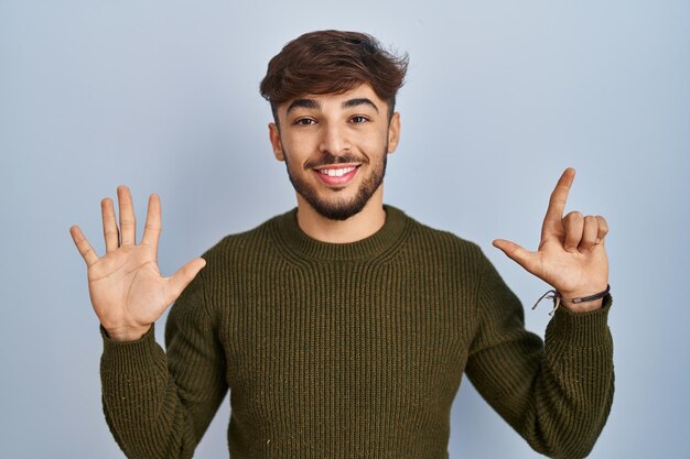 Arabski mężczyzna z brodą stojący na niebieskim tle, pokazujący i wskazujący palcami numer siedem, uśmiechając się pewnie i szczęśliwie.