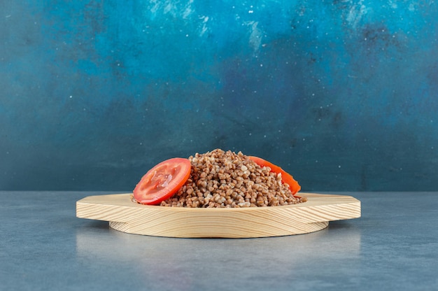 Aromatyczna Porcja Gotowanej Kaszy Gryczanej W Drewnianym Talerzu, Posypana Plastrami Pomidora, Na Niebieskim Tle. Zdjęcie Wysokiej Jakości