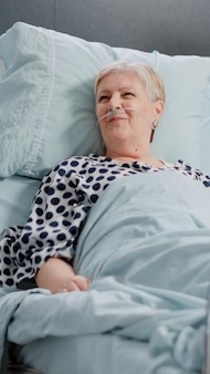 Asystent medyczny robi wizytę kontrolną starszego pacjenta w łóżku oddziału szpitalnego. kobieta pracująca jako pielęgniarka udzielająca pomocy choremu emerytowi z woreczkiem do kroplówki i donosowym przewodem tlenowym.